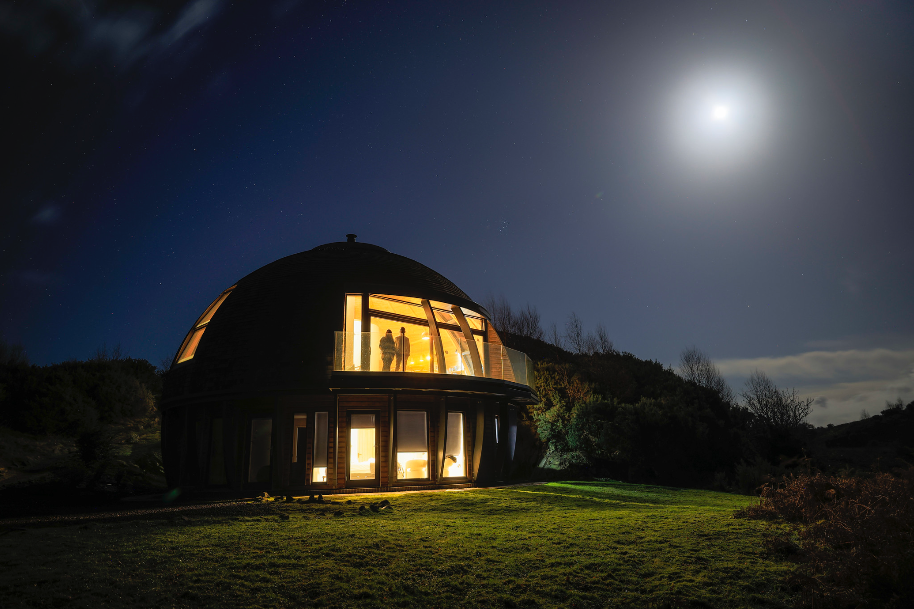 A couple stand on the balcony of a Loch Ken Eco Bothy under moonlight.